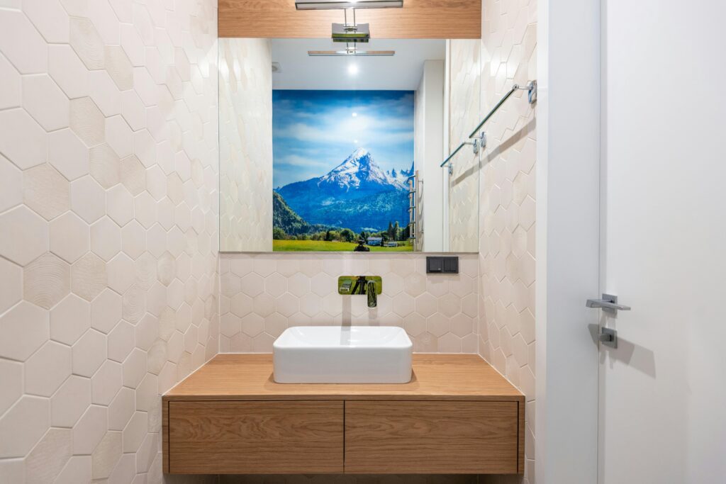 Wooden cabinet with white washbasin and tap placed near wall with mirror with reflection of wallpaper in contemporary washroom with door