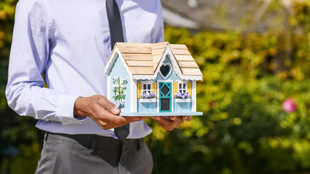 Businessman holds small house model outdoors, symbolizing real estate investment.
